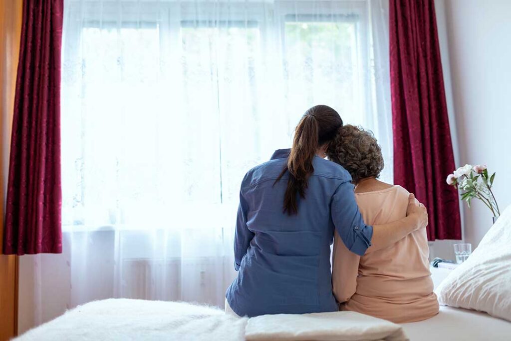 An elderly resident speaks with a caregiver in a warm, brightly lit common room.