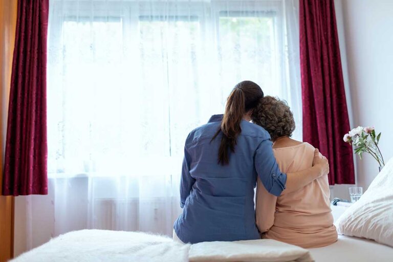 An elderly resident speaks with a caregiver in a warm, brightly lit common room.
